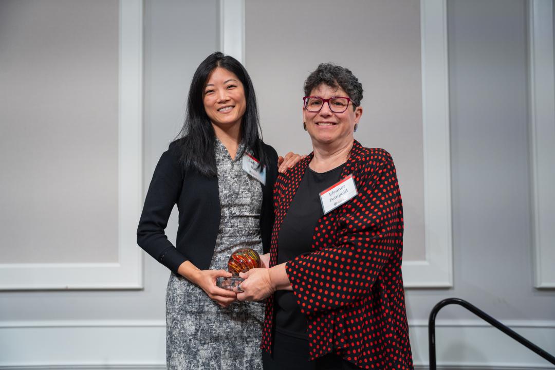 A woman in a black and white dress accepts an award from another woman in a black and red blazer.