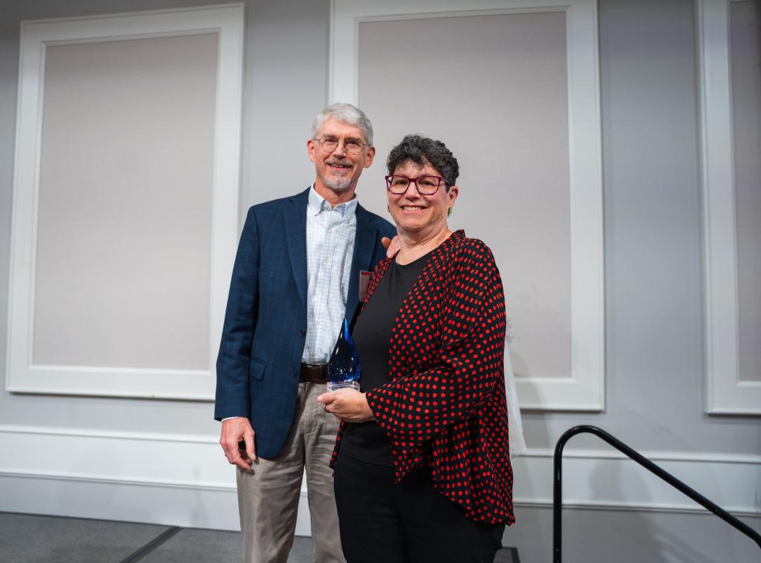 A man in a blue suit coat accepts an award from a woman in a red and black blazer.