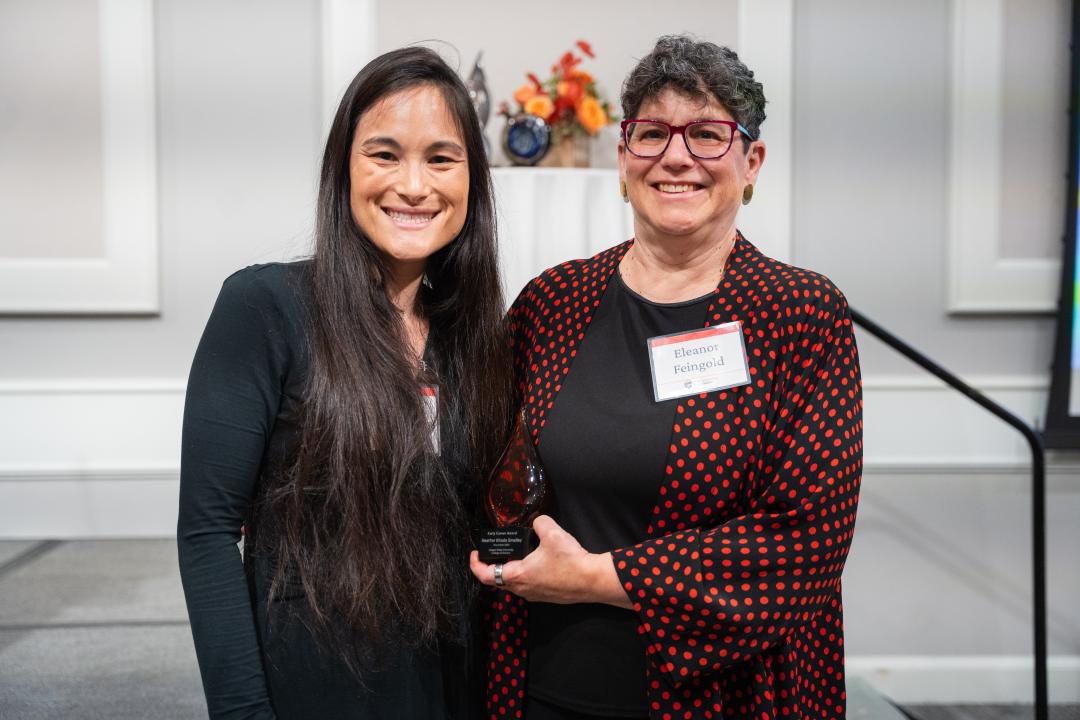 A woman in a black dress accepts an award from a woman in a black and red dress shirt.