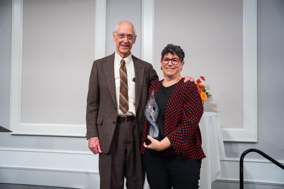 A man in a brown suit stands next to a woman wearing a red and black dress jacket. The woman is holing a glass award.