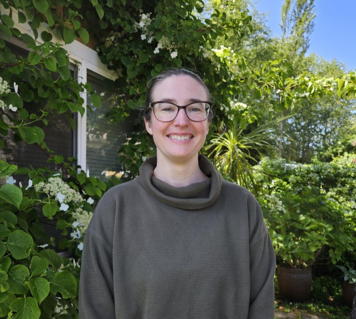 Mary Tunstall smiling for a photo under the sun in front of green leafy trees and bushes.