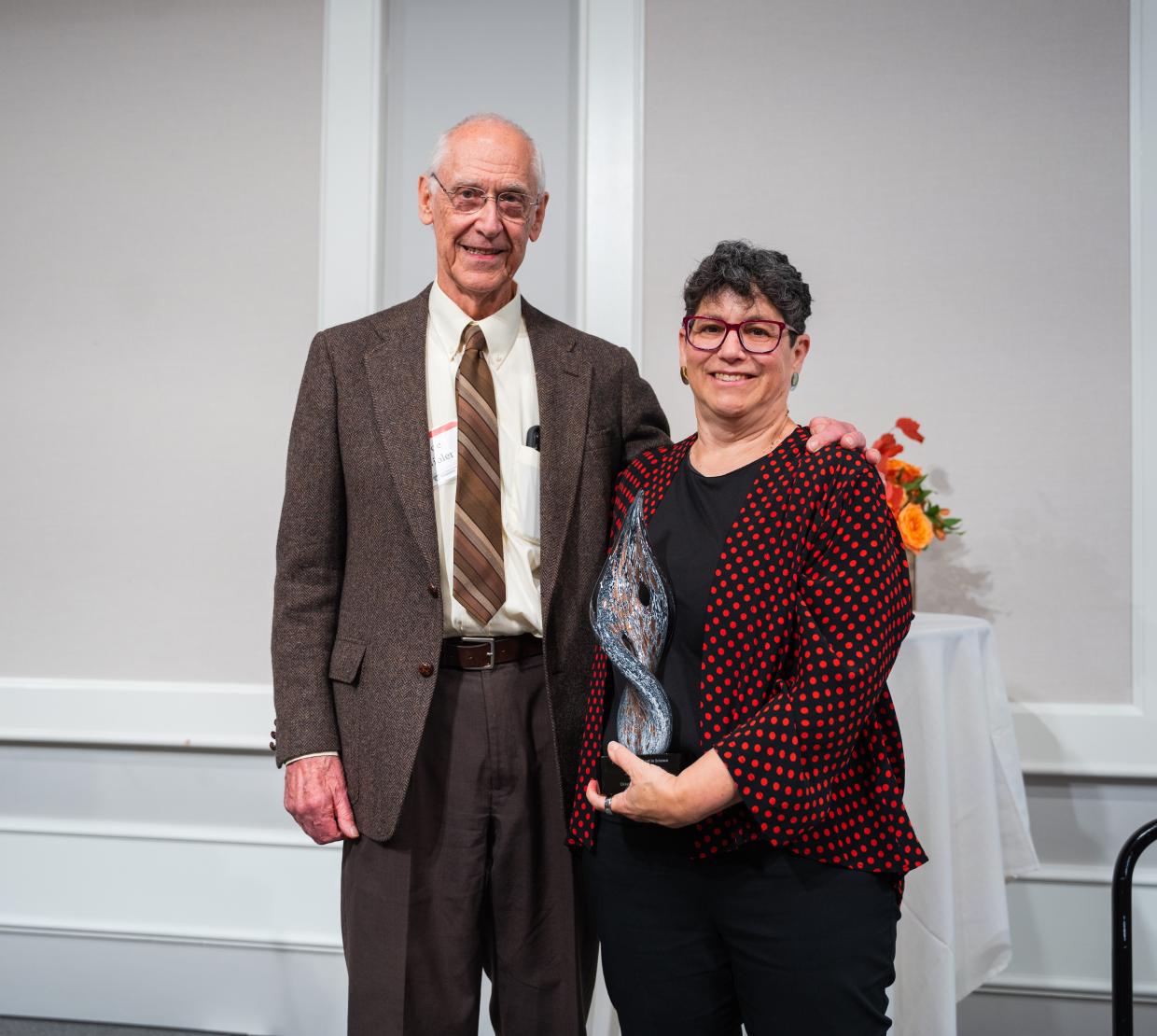 A man in a brown suit stands next to a woman wearing a red and black dress jacket. The woman is holing a glass award.