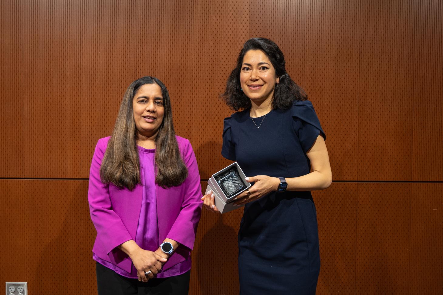 A woman in a purple shirt stands next to a woman in a black outfit holding an award.