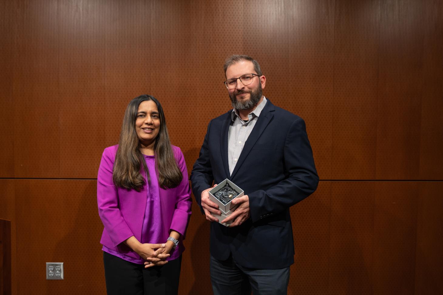 A woman in a purple shirt stands next to a man in a suit holding a glass award.