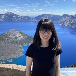 A woman in a black shirt poses for a photo in front of a stunning blue mountain lake.