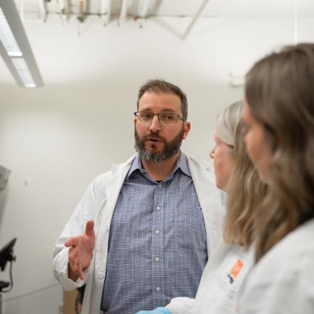 A bearded man wearing glasses and a button-up shirt beneath a lab coat speaks to two other scientists wearing lab coats toward the camera.