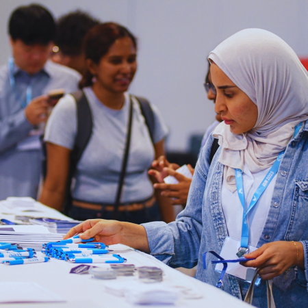 Woman wearing a hijab and a blue JSM lanyard peruses merchandise on a table with other conference attendees milling around.