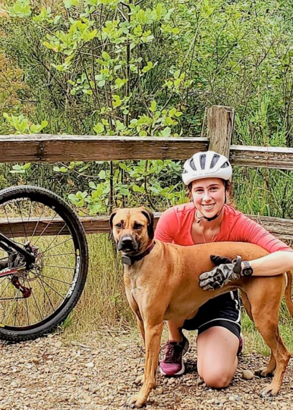 Smiling student standing with a dog and bike on an outdoor trail