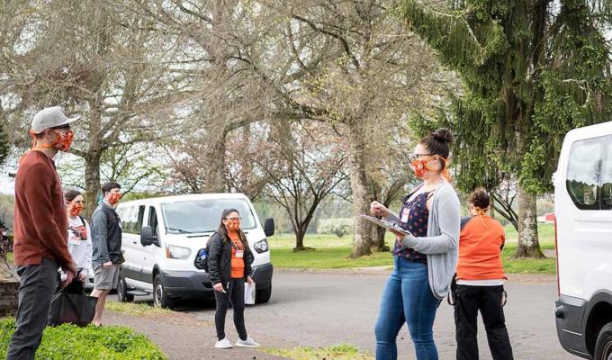 TRACE staff wearing face masks and face shields brief community volunteers before COVID-19 testing outside near two white vans.