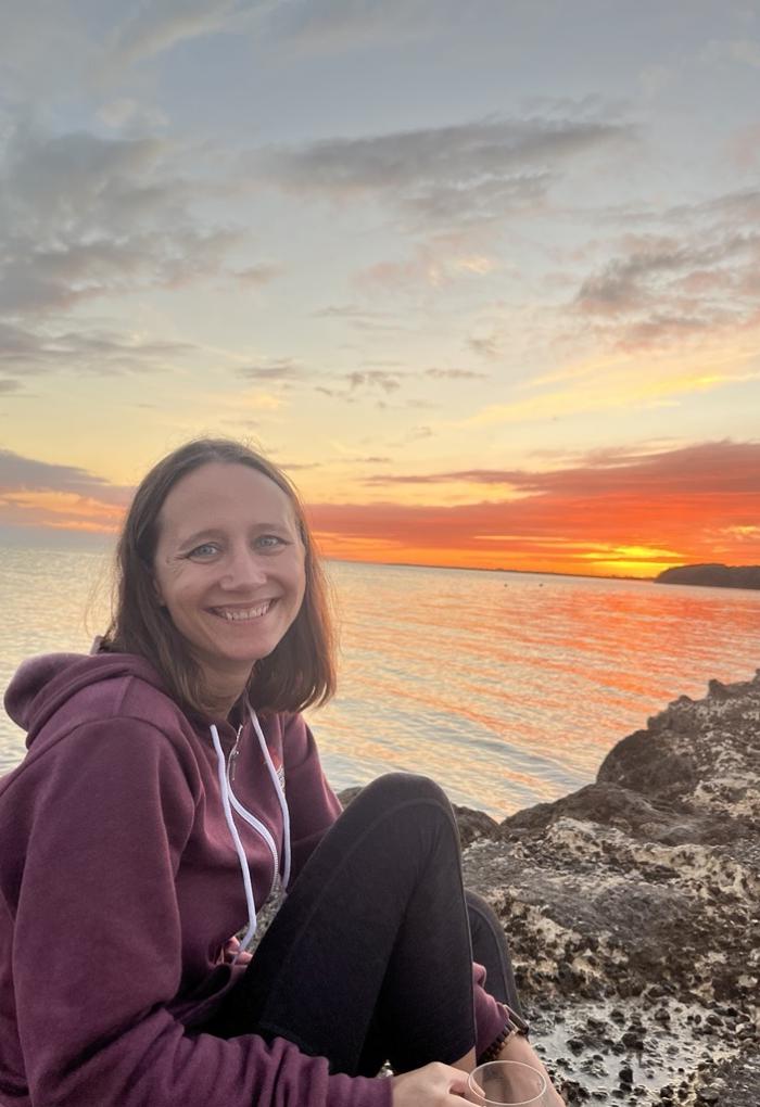 A woman poses for a photo in front of an orange sunset over water.