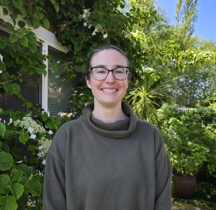 Mary Tunstall smiling for a photo under the sun in front of green leafy trees and bushes.