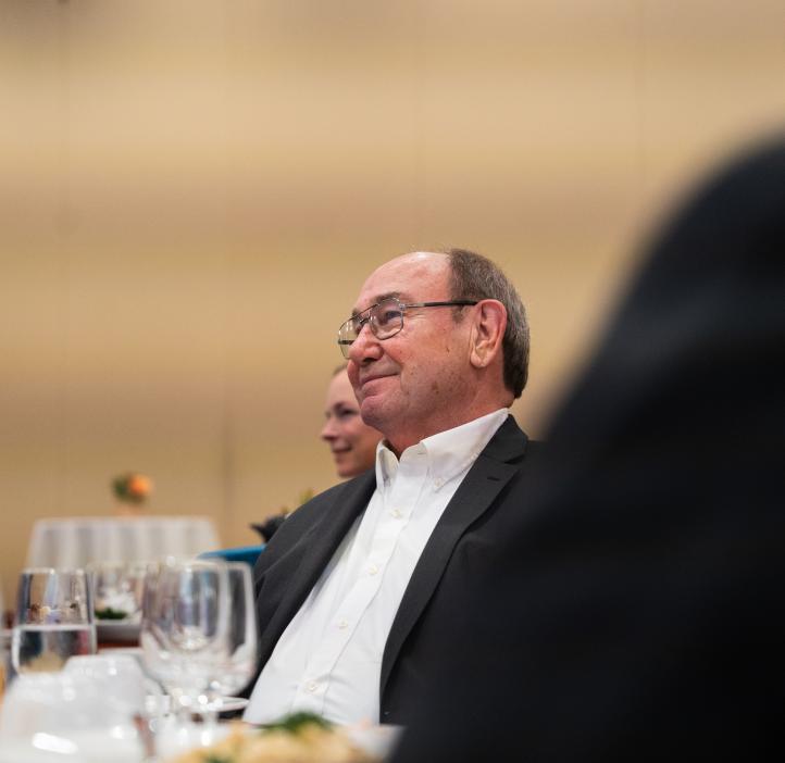 A man smiles fondly while looking off towards the left. He is sitting at a table wearing a suit jacket and white shirt.
