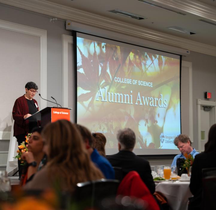 College of Science Alumni Awards Ceremony is in progress with a large screen displaying the event title. Attendees are seated at the tables. In a red patterned shirt, Dean Eleanor Feingold speaks at the podium.