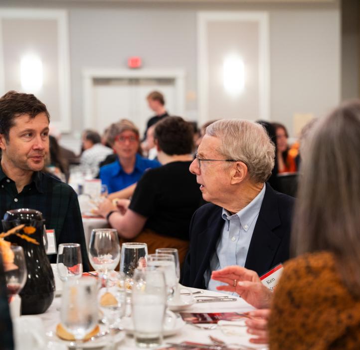Attendees seated at round tables engaged in conversation during the College of Science Alumni Awards dinner. The table is set with glasses, plates, and a pitcher. Other attendees are visible in the softly lit background. 