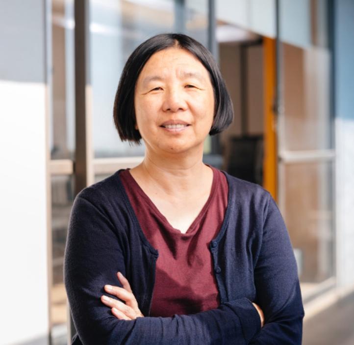 Headshot of Professor Bin Yu of UC Berkeley: woman in dark blue cardigan and maroon blouse, smiling with arms crossed while standing in front of college building with large glass windows.
